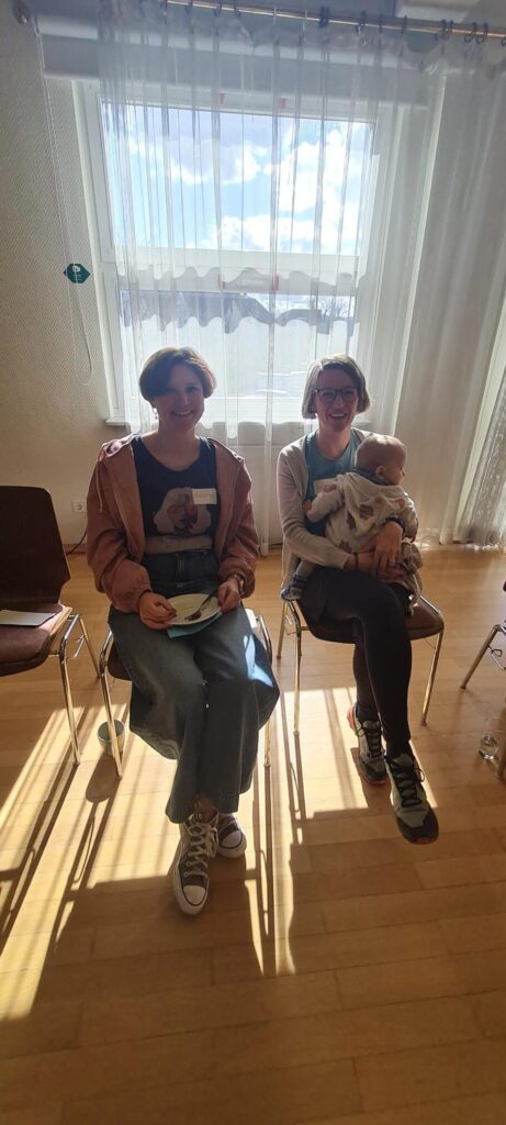 Zwei Seminar Teilnehmende auf Stühlen sitzend. Eine mit Teller und eine mit Baby auf dem Schoß.