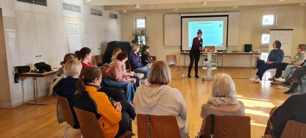 Seminar Teilnehmende im Halbkreis auf Stühlen. Davor eine Person, die eine Präsentation zeigt.